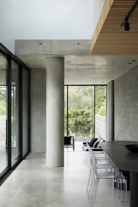 Modern minimalist residential interior with concrete walls and floors, large windows showing natural light, and a sleek dinin