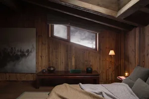 A cozy, rustic bedroom with wooden walls, a bed with soft bedding, and a small table with a lamp, candle, and painting.