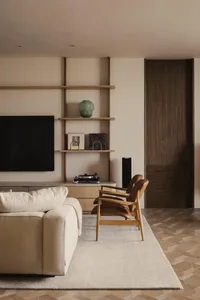 A modern minimalist living room with a beige sofa, brown armchairs, and a wooden shelf with books and a vase.