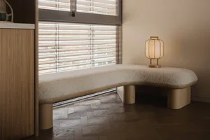 A modern bedroom featuring a wooden bench with a light-colored textured cushion placed next to a large window with blinds.