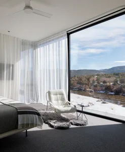 Modern minimalist bedroom featuring a white leather chair with a glass window overlooking mountains and a river.