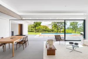 A modern living room featuring wooden tables, chairs, and a glass coffee table with a view of a pool and greenery.