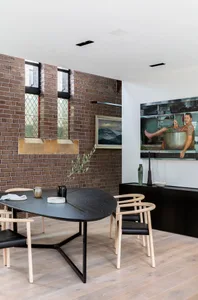 A modern dining room with a black wood table and chairs, set against a brick wall with stained glass windows.