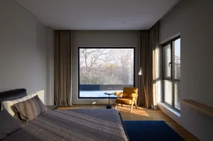 A modern bedroom featuring gray flooring and a blue rug, with natural light streaming through large windows and a sitting are