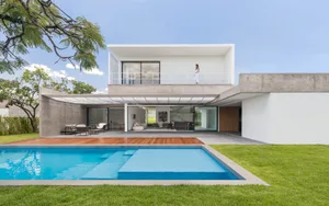 A modern house with a concrete exterior and a glass balcony overlooks a peaceful lawn and swimming pool.
