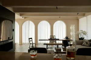 A modern living room with cream-colored walls and arched windows, featuring a wooden table, chairs, and a sofa with a glass a