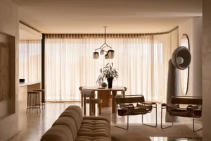 A modern living room with neutral-toned furnishings, a round dining table with hanging lamps, and large windows with light-co