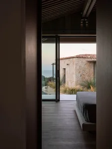 A modern bedroom with wooden flooring, a gray bed, and a view of a house with a red-tiled roof and sea in the background.