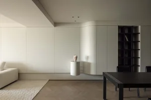 Modern minimalist living room featuring white walls, furniture, and floor, with a vase on a side table and a dark wooden tabl