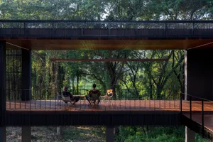 Three people sitting on a wooden deck surrounded by trees under modern industrial architecture.