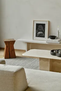 A minimalist living room with a white sofa, wooden stool, marble coffee table, and abstract art on the wall.