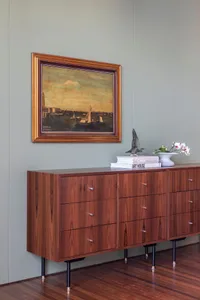 A modern, wooden sideboard with a painting on the wall, books, and a vase with flowers.