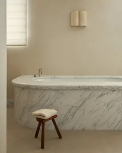 Modern minimalist bathroom featuring a white marble bathtub, a wooden stool with a folded towel, and a sconce on the wall.