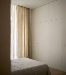 Modern minimalist bedroom featuring a bed with white sheets and curtains made of light-colored fabric, against a wall with a 