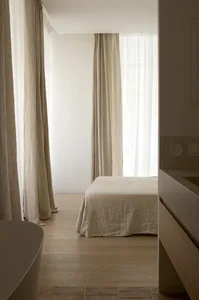 A minimalist bedroom with neutral-toned curtains, a single bed, and wooden flooring.