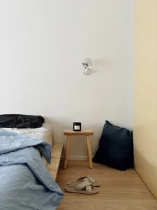 Minimalist bedroom with a bed, stool, and slippers on a wooden floor, featuring a wall-mounted light fixture.