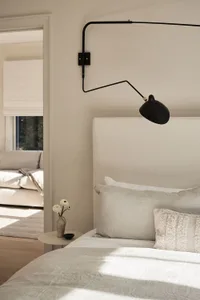 A minimalist bedroom with a white bed and light-colored textiles, featuring a sleek black wall lamp.