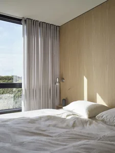 A minimalist bedroom with a textured wooden wall and a grey bed against a large window.