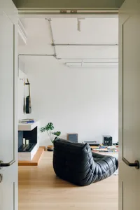A minimalist living room with a black leather sofa, a wooden floor, and a white wall.