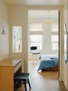 A modern, minimalist bedroom featuring hardwood flooring, a blue bedspread, and a dark blue upholstered stool.