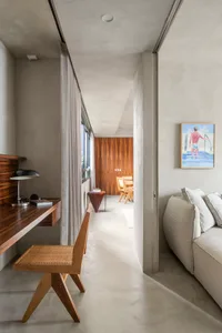 A modern minimalist interior space with concrete walls and floors, featuring a desk, chair, sofa, and artwork.