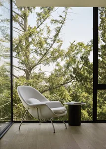 A minimalist living room with a white upholstered armchair, black metal side table, and wooden flooring, featuring a view of 