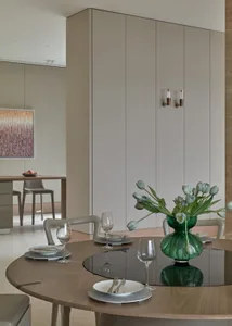 A modern dining room featuring a wooden table with plates, cutlery, and glasses, accompanied by a green vase with tulips on a