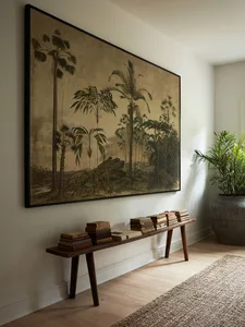 A photograph of a living room featuring a large framed painting of tropical foliage, a wooden bench adorned with stacked book