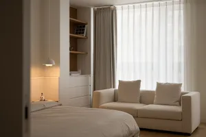 A minimalist bedroom featuring a comfortable sofa with white cushions, white curtains, and a white pendant light.