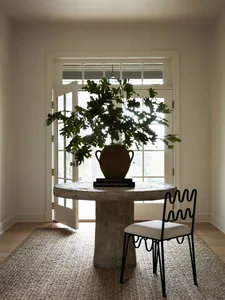 A minimalist interior featuring a round concrete table with a plant and a single chair on a textured rug, in front of a large