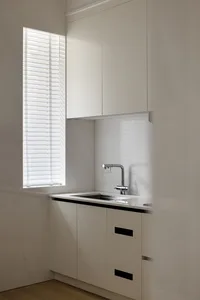 A modern, minimalist residential kitchen with white cabinets, a sink, and a window with blinds.