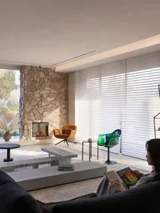 A modern living room featuring a stone-clad fireplace, minimalist furniture, and large windows with white blinds.