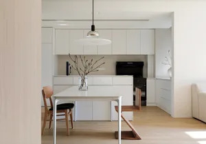 A modern, minimalist kitchen with white cabinetry and a wooden dining table.