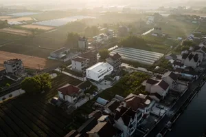Aerial view of a village with houses, fields, and greenhouses under a hazy sky.