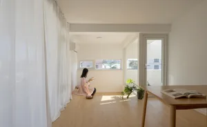 A minimalist living room with wooden flooring and a person reading a book by a window.