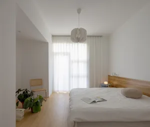 A minimalist bedroom with white bedding, wood flooring, and white lighting fixtures.