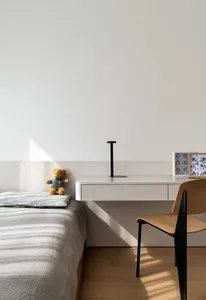 A minimalist bedroom featuring a bed with a grey blanket, a small shelf with a teddy bear and a black lamp, and a wooden chai