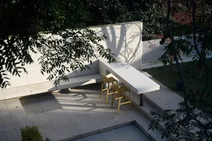 A modern outdoor space featuring a white concrete seating area with a table and stools, surrounded by lush greenery and trees