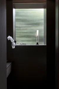 A minimalist bathroom featuring a frosted glass window with a sleek, modern lamp on the sill and a white orchid on the left s