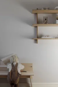 A child sitting at a minimalist desk with a wooden shelf overhead containing books and toys.