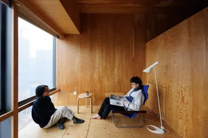 A minimalist living space with wooden walls and floor, featuring two individuals sitting and relaxing.