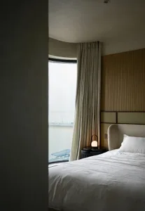 An elegant minimalist bedroom featuring beige curtains, a beige headboard, and a white lighted lamp on a bedside table, with