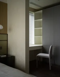 A modern minimalist bedroom featuring a bed with white sheets, a wooden headboard, a built-in desk with shelves, and a white