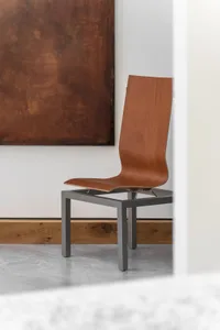 A minimalist wood chair with a sleek design, sitting in front of a brown abstract painting on a white wall.