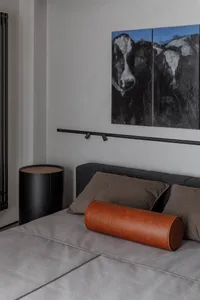 Modern bedroom with a gray bed, leather cylindrical pillow, and cushions, featuring abstract paintings of cows on the wall.