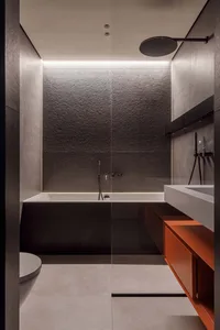 Modern, minimalist bathroom with concrete walls, a floating vanity with orange accents, and a sleek white bathtub.