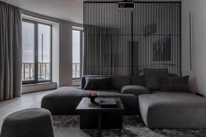 A modern, minimalist living room with a grey sofa, wooden floor, and large windows.