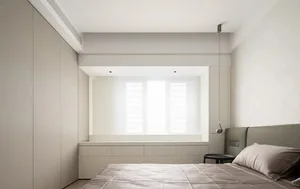 Neutral minimalist bedroom featuring a bed with a simple grey headboard, a window with white shutters, and built-in storage u