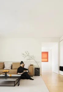 A minimalist living room with neutral tones, featuring a person sitting on a brown sofa, reading a magazine, and placing oran