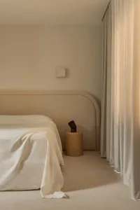 A minimalist bedroom featuring a bed with a textured cotton blanket, neutral-colored curtains, and a simple side table with a